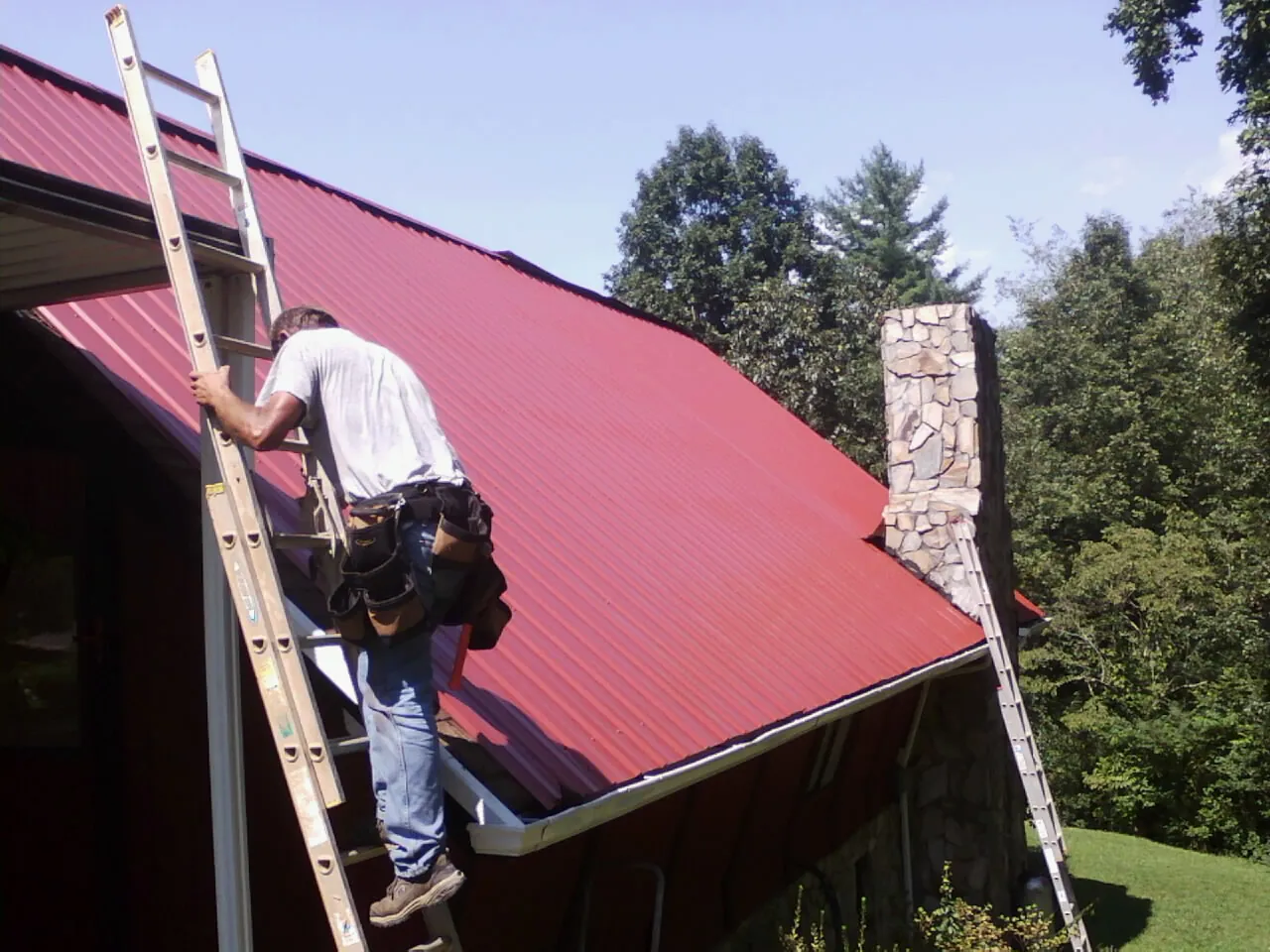 Roofer on a ladder beside a red metal roof and stone chimney — Soffit & Fascia Repair by Roofing Brothers in Marlboro